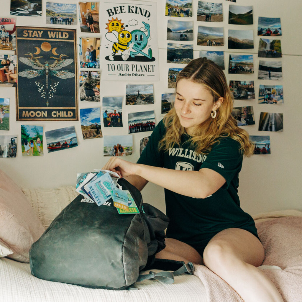 student on bed with backpack and photos on wall